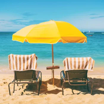 two beach chairs with towels under a yellow umbrella on a beach in a sunny vacation destination