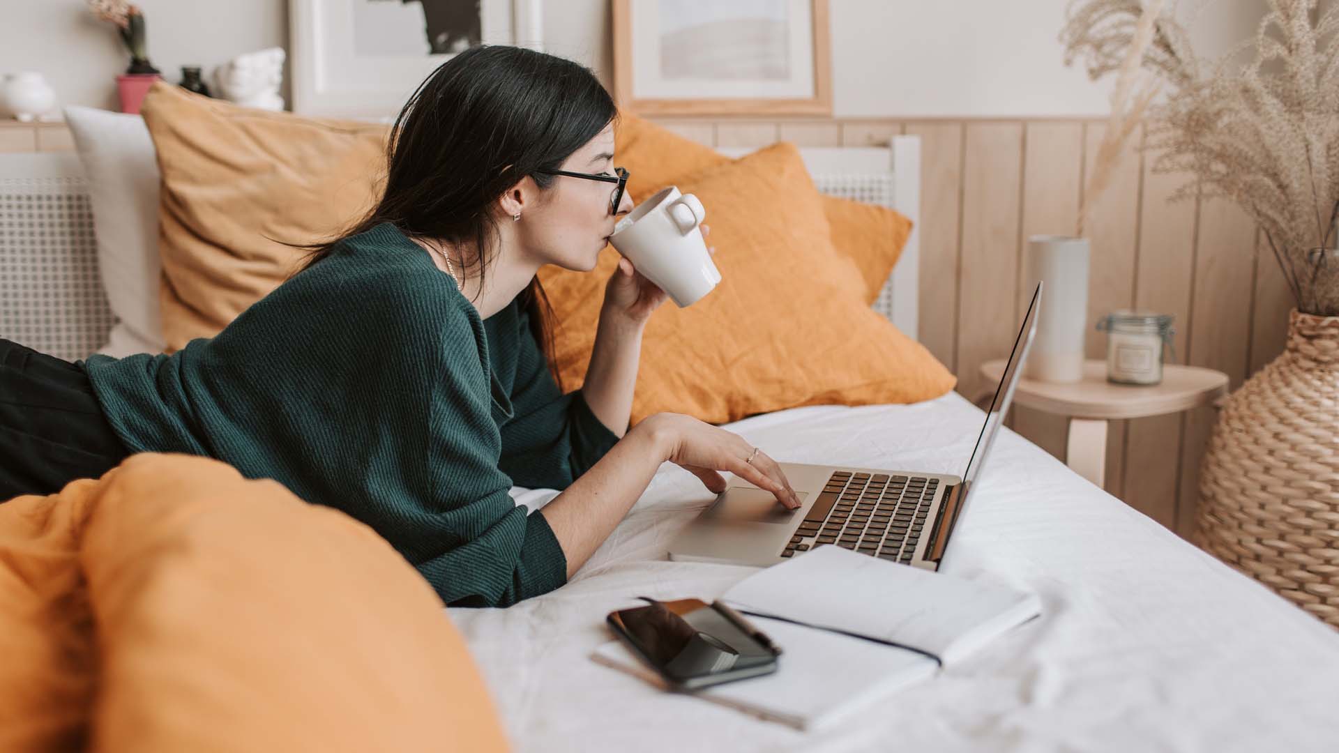 Woman on bed on laptop and drinking coffee