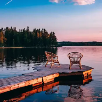 Two chair at the end of a dock on a lake at sunset.