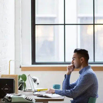 Man sitting at desk in front of laptop next to window.