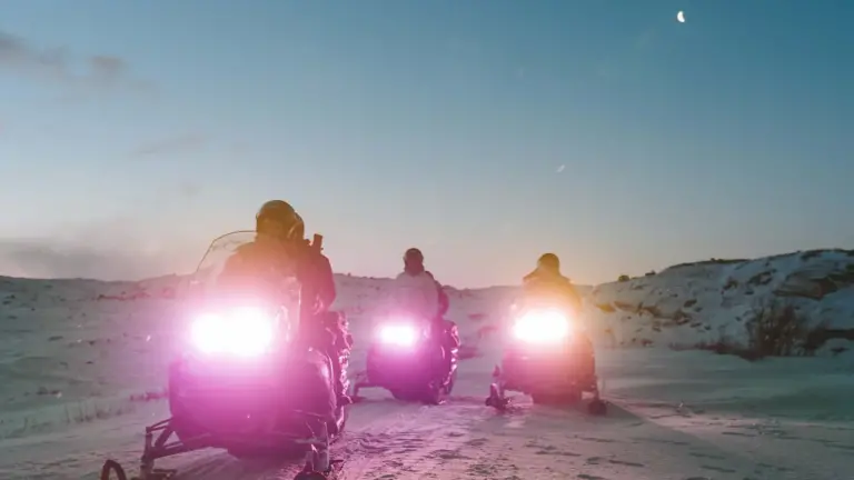 Three snowmobiles driving with their lights on at nighttime.